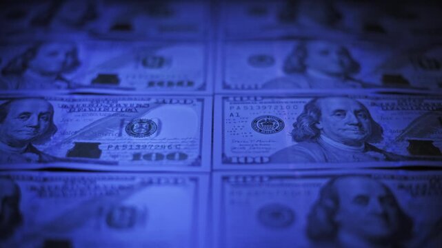 Low angle close-up rack focus moves across rows of one hundred dollar bills under a blue ultraviolet light. The authenticity check reveals security features on US currency in a dark setting.