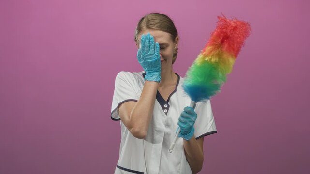 Cleaning woman in white uniform holds rainbow feather duster and covers face with gloved hand against pink studio backdrop; fatigue.
