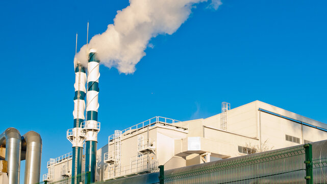 A heating plant and pipes emitting white smoke, with a pipeline in the foreground and a blue sky in the background
