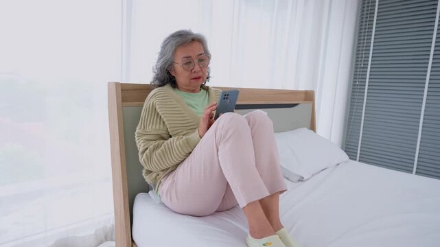 Modern Senior Using Smartphone in Bed: A composed senior woman sits comfortably on a bed, deeply engaged with her phone, embodying a sense of relaxation and connection.