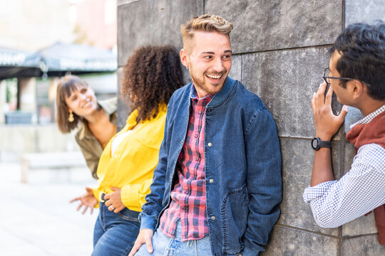 Playful young friends laughing around a city wall during a casual outdoor break. Multiethnic millennials and gen z share jokes in an urban hangout. Candid lifestyle concept