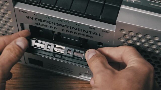 High-angle close-up of hands inserting a vintage audio cassette into a silver boombox. The door is closed, and the play button is pressed to start music on a wooden surface.