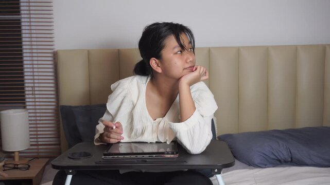 Teenage girl using tablet on bed at night while stressed thinking. Asian student hold stylus pen to study online inside bedroom during Gen Z lifestyle homework exam preparation.