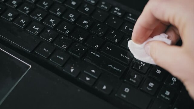 Close-up of a hand spraying antiseptic liquid onto a dusty black laptop keyboard and wiping it clean. The action highlights hygiene practices and the importance of sanitizing shared devices.