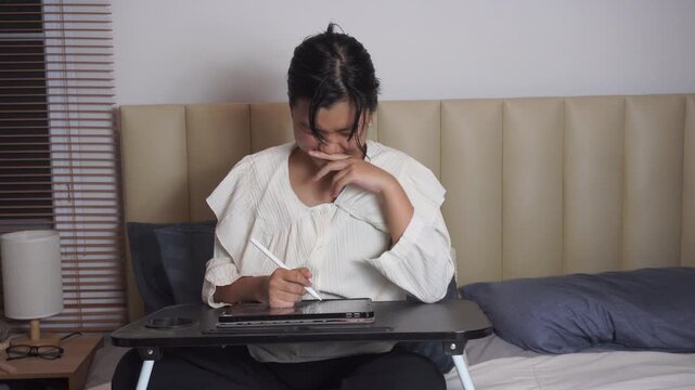 Teenage girl using tablet on bed at night. happy Asian student is drawing with stylus pen inside bedroom during her Gen Z lifestyle creative art hobby remote freelance job