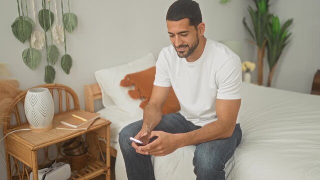 Man sitting on bed holding smartphone and smiling, wearing white tee and jeans beside rattan nightstand with lamp, notebook and plants in bedroom; relaxed ease.