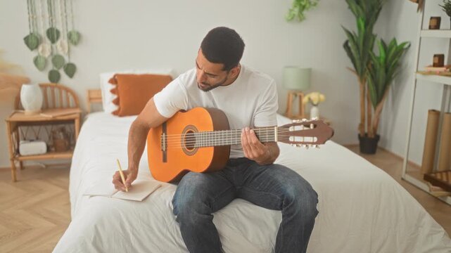 Man playing an acoustic guitar on a bed with hands fretting strings in a studio space, jotting lyrics in a notebook beside him; songwriting calm.