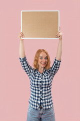 Young woman with red hair and a cheerful expression holding up a blank rectangular sign or board...