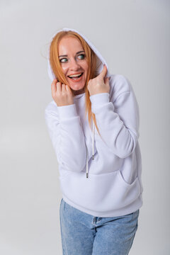 Young woman with red hair and green eyes expressing happiness and excitement, holding her white hoodie playfully while looking away on a studio background