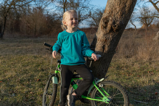 Blonde girl leaning against a tree with a green bicycle in a sunny grassy field, smiling and enjoying outdoor play, childhood freedom and simple nature moments