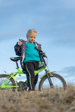 Young girl standing with bright green bicycle on a dry grassy hill, adjusting her jacket and looking down while enjoying a sunny outdoor biking moment in nature
