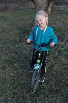 Blond girl in a blue sweatshirt and black pants happily stands with her children's bicycle on green grass, enjoying playful outdoor learning and carefree childhood balance