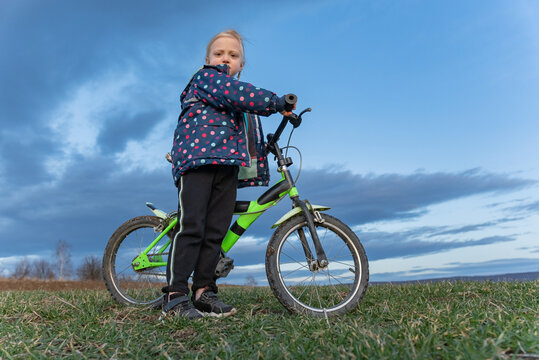 Young girl standing outdoors with a green and black bicycle on a grassy field, enjoying childhood and outdoor activity under a cloudy sky, representing learning and growth