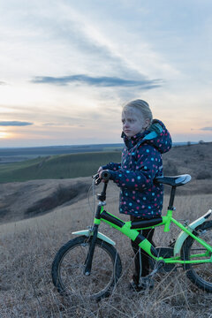 Young blonde girl in a polka dot jacket stands on a dry grassy hill at sunset, holding a bright green bicycle and gazing pensively over a quiet rural landscape under cloudy skies