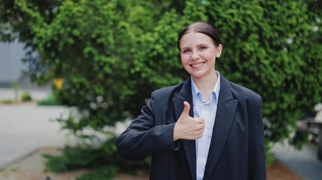 Professional woman smiles happily, giving approval with thumbs up gesture. Conveys confidence, success, and positive outlook outdoors. Represents well-being, achievement, and excellent performance.
