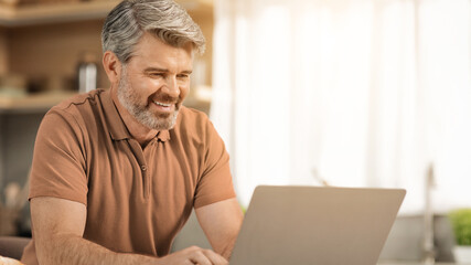 Fototapeta premium Smiling Elderly Caucasian Man Working On Laptop At Home Office, Engaged And Focused While Typing At Desk With Natural Light. Business Concept For Remote Work And Senior Professionals.