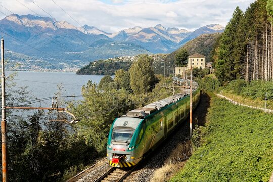A train of Trenord Railway traveling along Lake Como on a sunny summer day with alpine mountains in background and clouds in the blue sky, in Dorio, Lecco, Lombardy Region, Italy