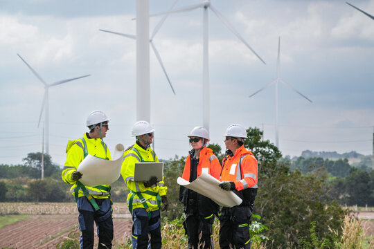 A four person engineering crew in yellow and orange hi vis walk the boundary while reading the site plan crossing farmland inside an operating wind park in bright cloudy daylight at a wind.