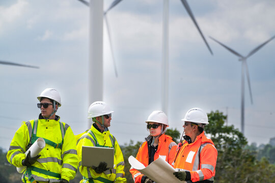 A four person engineering crew in yellow and orange hi vis pause to argue over an open blueprint crossing farmland inside an operating wind park in bright cloudy daylight on a working wind.