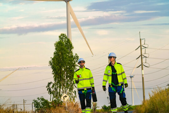 Two technicians with a tool case talk quietly on the dusk lit straight on an empty rural road through the park in hazy sunset light at an operating renewable power project.