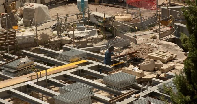 An overhead shot reveals an active archaeological excavation site, featuring a complex network of metal structural supports, scaffolding, and stone foundations where researchers carefully work.