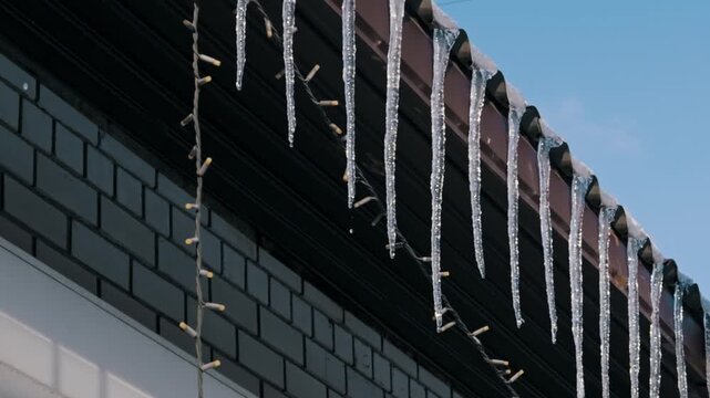 Long, clear icicles hang from a roof eave, dripping water as they melt in the sun. The camera slowly pulls back against a backdrop of a clear blue sky and dark wall.