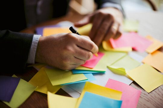 Male office worker in a suit writing on a sticky note surrounded by colorful notes on a wooden desk, emphasizing organization and brainstorming activities