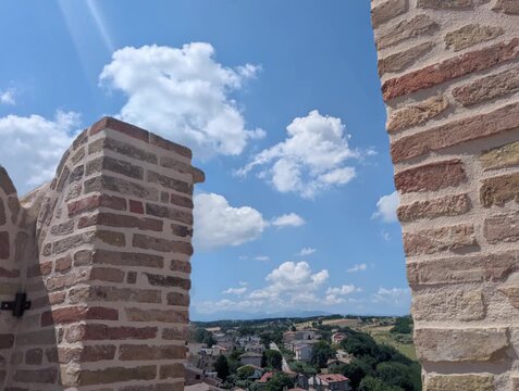 Un muro in mattoni emerge in primo piano sotto un cielo limpido, mentre sullo sfondo si apre un paesaggio di colline e piccoli edifici
