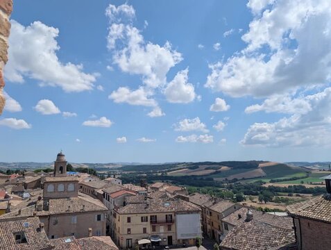 Il borgo storico si apre con tetti in cotto e edifici in pietra che dominano la scena, mentre sullo sfondo le colline verdi marchigiane creano un panorama luminoso e armonioso