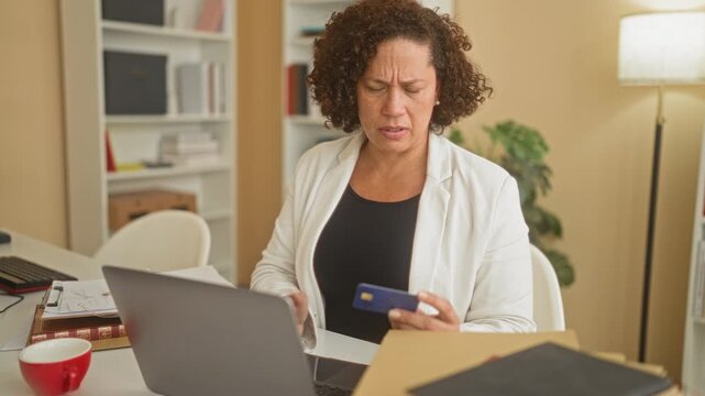 Woman holding creditcard while using laptop in building office, furrowed brow and frowning with card in hand; financial concern.