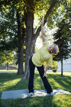 Senior woman in yellow shirt performing a side stretch exercise on a yoga mat in a park with trees and grass surrounding her in bright daylight