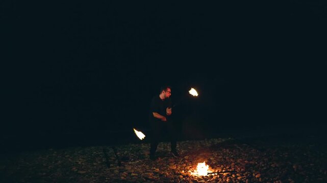 Man performing a fire show with torches on a pebble beach at night.