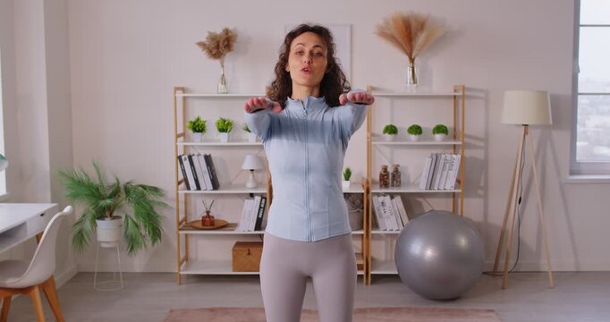 Slim woman sport coach doing physical exercise while streaming online to followers, raising legs to hands, demonstrating how to stay healthy and fit, dressed in activewear, posing indoor