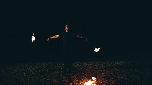 Man performing a fire show with torches on a pebble beach at night.
