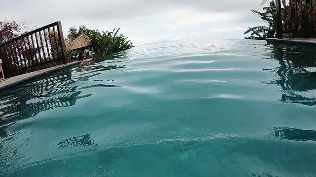 First person pool view shows gentle surface ripples and brighter foreground water movement, with tropical resort railings, lounge furniture, lush greenery, palm leaves, and calm horizon under soft