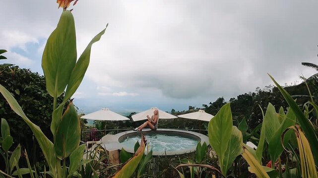 Seated woman in swimwear relaxes beside circular resort pool, framed by dense tropical plants, white parasols, and distant hillside landscape under soft overcast daylight, viewed from low angle with
