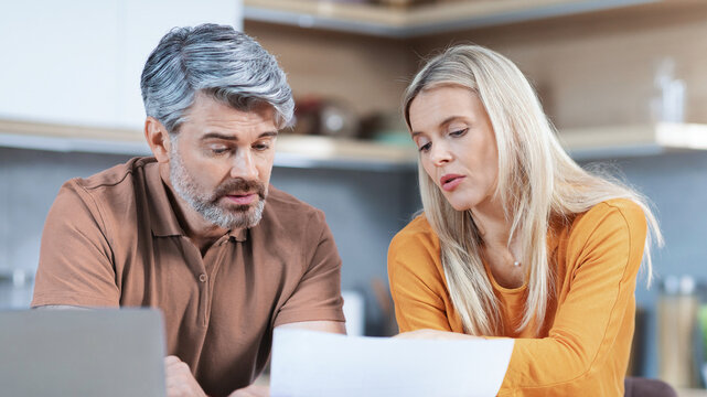 Worried Middle Aged Caucasian Man And Woman Looking At Papers Together While Sitting At Home, Engaged In Discussion About Important Documents On Table. Business Concept.