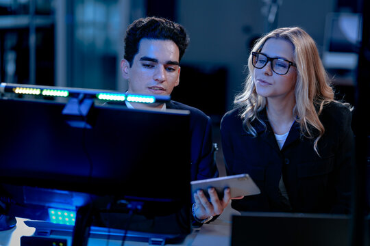 Intense late night project review. Female team leader points out critical data on screen discussing report charts with colleagues in modern high tech operations center.