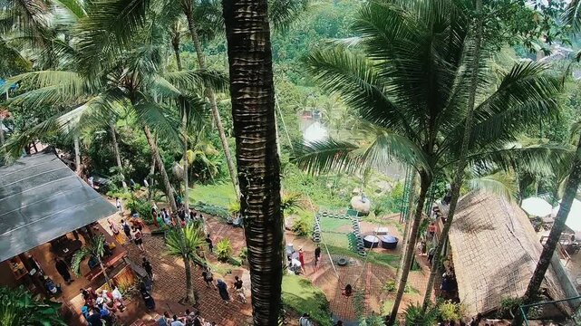 Elevated view over crowded tropical resort terraces with visitors moving along brick walkways beside thatched shelters, outdoor seating, dense palms, and layered green hillside scenery in bright
