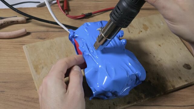 Hands use a professional heat gun to carefully shrink blue protective wrap around a custom battery pack on a wooden workbench. Concept of DIY electronic repair and manufacturing process.