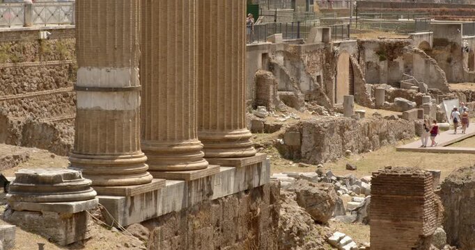 Closeup of three massive, fluted ancient Roman columns stand on a stone platform in the foreground, framing a vast archaeological excavation site filled with crumbling stone structures and ruins.