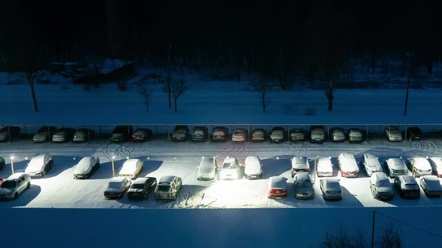 Parked vehicles sit in a snow-covered lot at night while vivid traffic light trails streak rapidly across the adjacent road. Concept of winter commuting and the relentless passage of time.