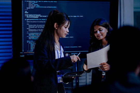 An Indian female developer leads a code review, holding a report. She explains a Python script for an AI or cybersecurity system to her team in a dark SOC Security Operations Center