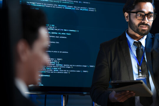 An Indian male software engineer or team lead discusses Python code for an AI algorithm. A developer in the audience raises their hand to ask a technical question during a code review.