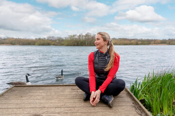Fototapeta premium Woman sits on dock by water with geese while enjoying the outdoors on a sunny day in a park