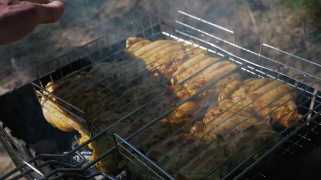 Marinated chicken steaks are grilling on a metal wire rack over hot burning charcoal. Wisps of smoke rise from the juicy meat as it cooks to a golden brown perfection in the outdoors.