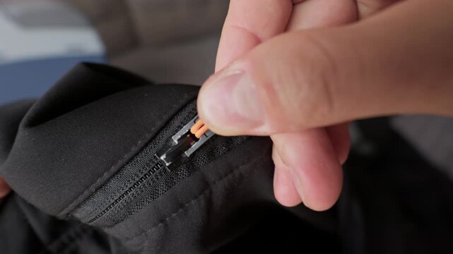 Male fingers firmly pull a bright orange zipper cord to fasten a black outdoor jacket. Concept of getting ready for cold weather, securing camping gear, and functional clothing details.