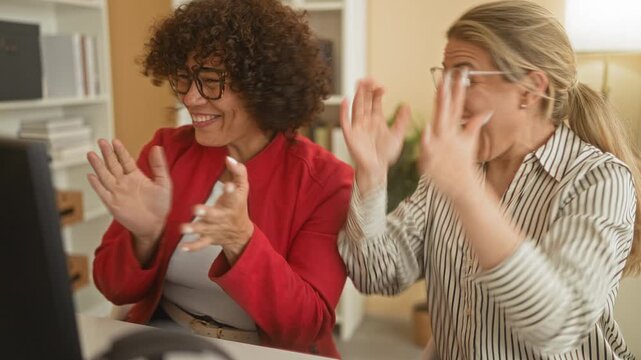 Woman in red jacket clapping hands at desk in building office, smiling with raised fists while colleague applauds beside her; team success joy.
