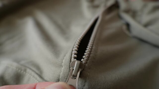A close-up view shows fingers pulling a plastic zipper pull on an olive green textile garment or jacket. Concept of garment construction, getting dressed, and textile manufacturing.