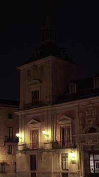 Vertical night view of an illuminated historic Spanish government building with classical stone architecture, decorative facade, balconies, and warm lighting against a dark sky.